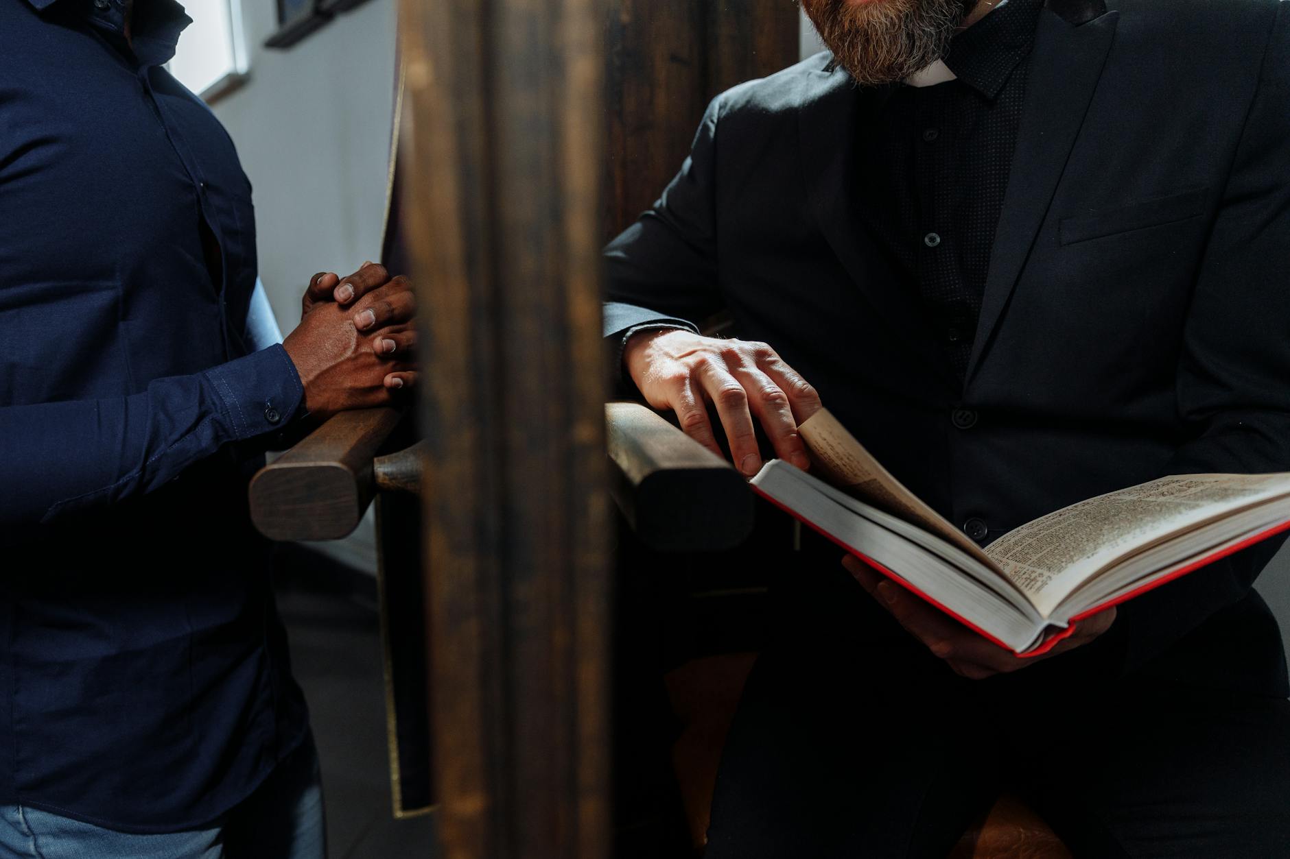 priest holding bible behind wooden wall while man confessing